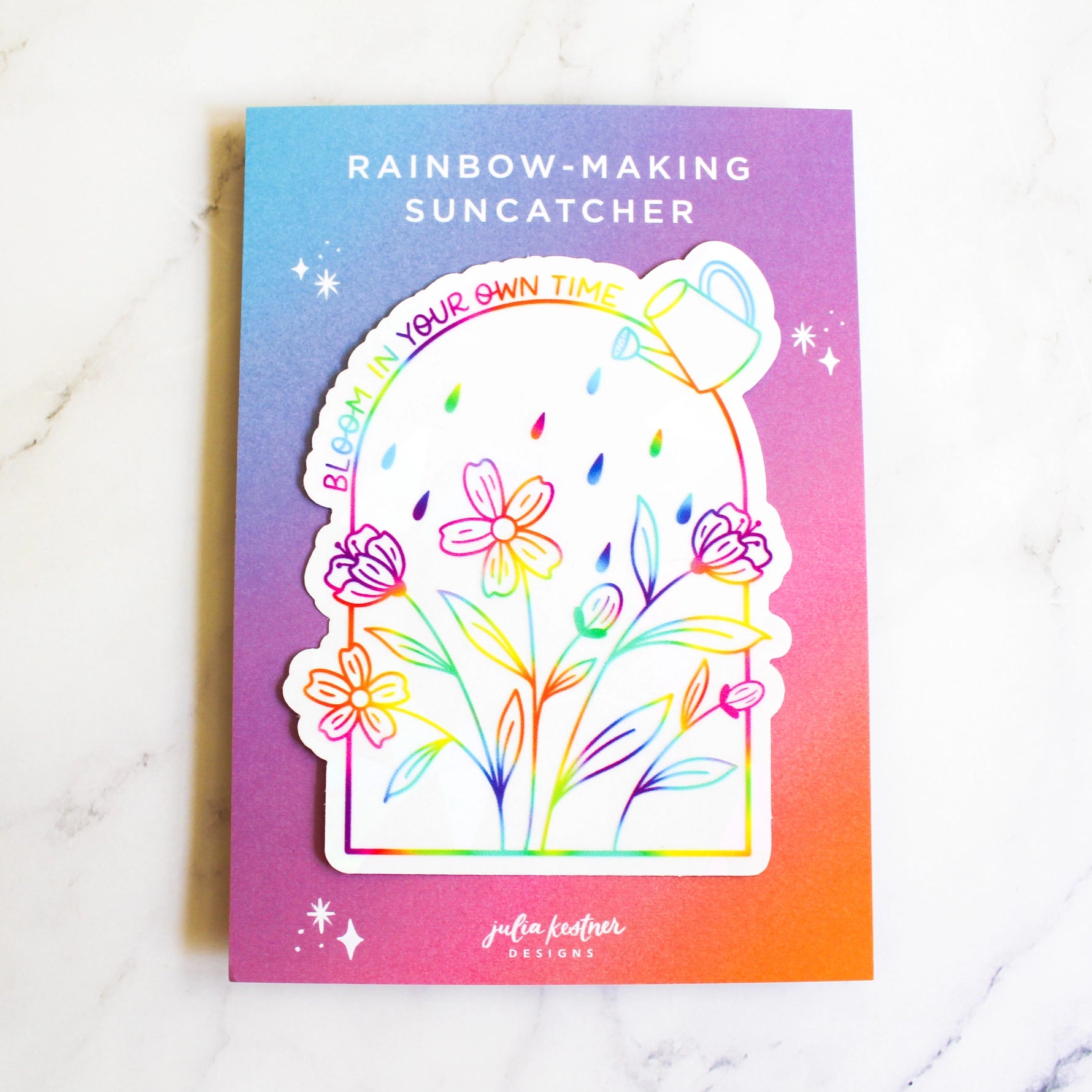 A colorful card or sticker set titled "RAINBOW-MAKING SUNCATCHER" on a white marble surface. The design features a white die-cut sticker with rainbow-lined flowers, leaves, rain drops, and a watering can, placed on a purple-to-orange gradient backing card with the text "BLOOM IN YOUR OWN TIME" and the brand name "julia feather designs" at the bottom.