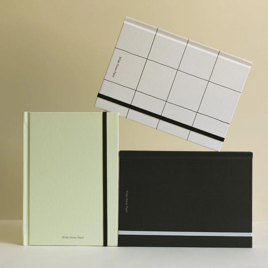 Three hardcover notebooks arranged on a white surface against a warm beige background. One notebook is pale mint green with a black elastic band, one is black with a white horizontal stripe, and one is white with a black grid pattern and black stripe.