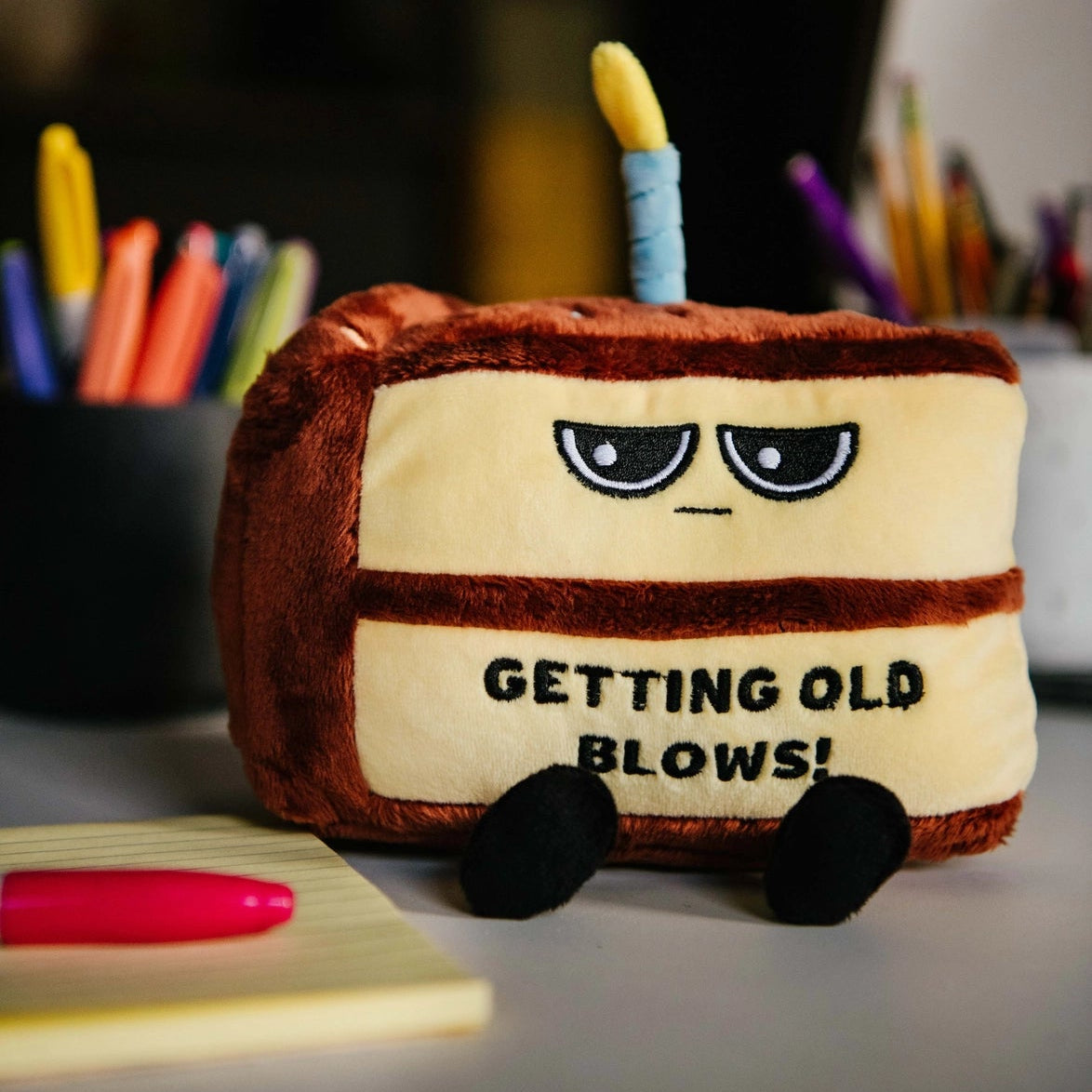 Plush toy resembling a birthday cake with a candle and text 'GETTING OLD BLOWS!' on a desk with stationery items.