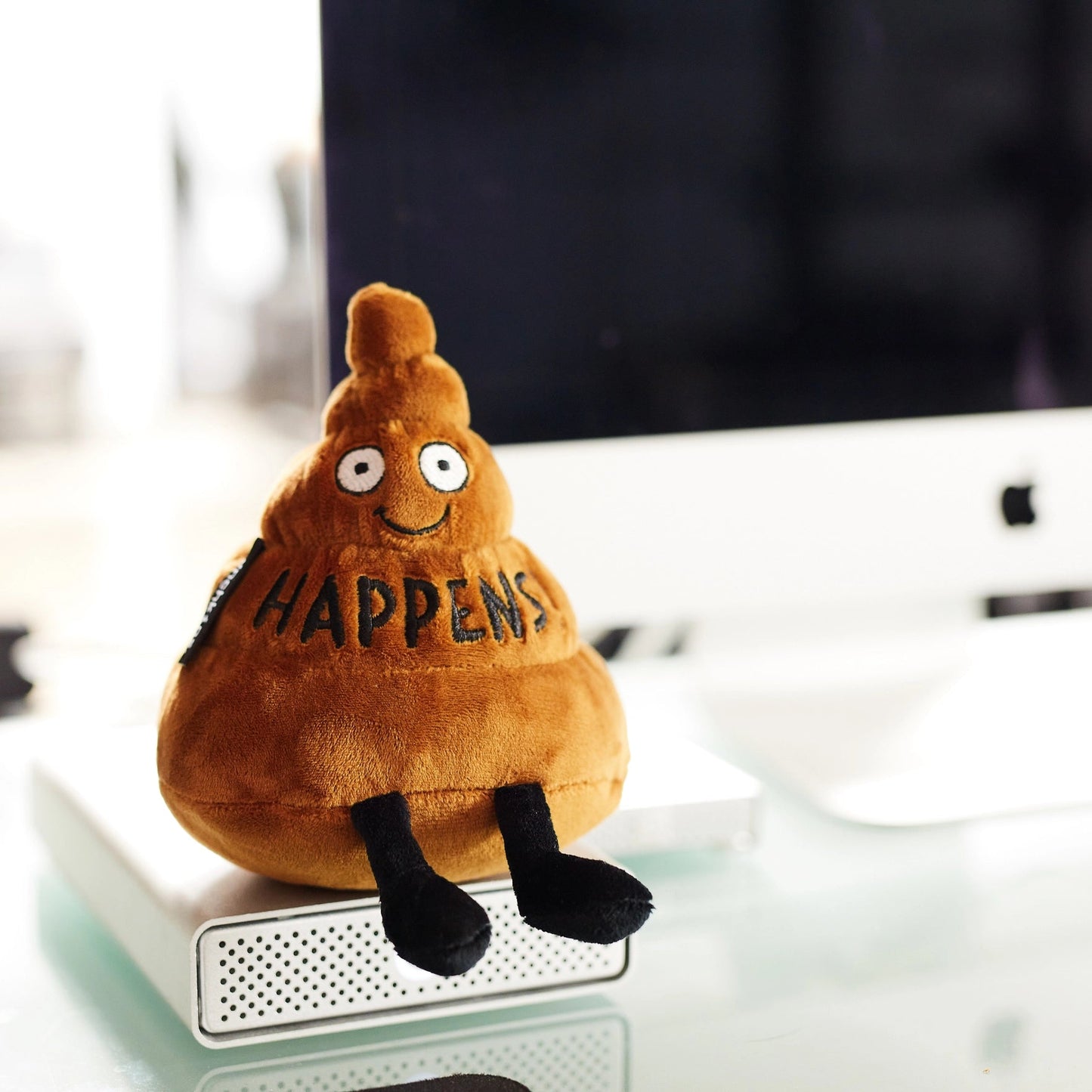 Plush toy resembling a poop emoji with 'HAPPENS' text on a desk.