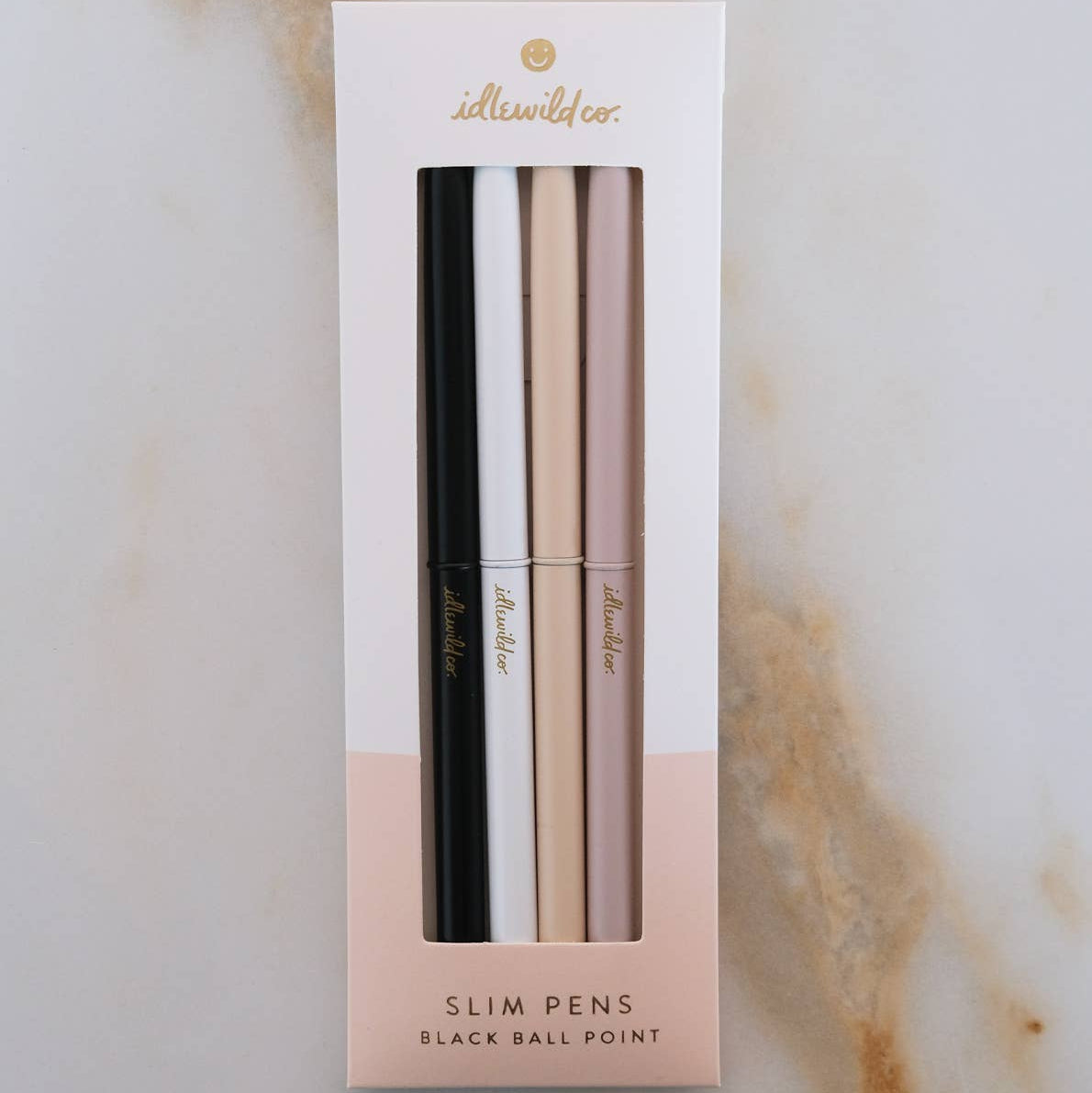 Set of four slim pens in black, white, cream, and rose gold packaging on a marble background